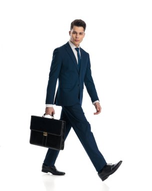 Bavullu, lacivert takımlı seksi genç adam ileriye bakıyor ve beyaz arka planda yürüyor.