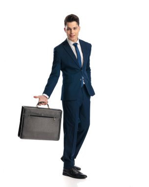 Lacivert takımlı yakışıklı genç adam gülümsüyor ve beyaz arka planda bavulu tutuyor.