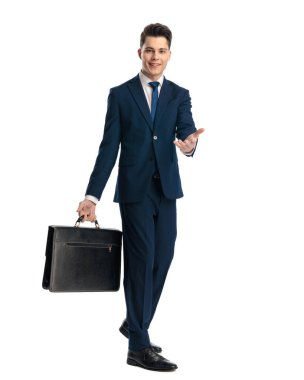 Lacivert takım elbiseli mutlu bir işadamı elinde bavuluyla beyaz arka planda gülümserken elini ileri uzatıyor.