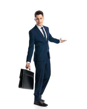 Kendine güvenen mavi takım elbiseli, yanında bavulu olan ve beyaz arka planda gülümseyen genç bir adamın yan görüntüsü.