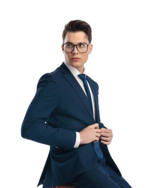 Gözlüklü genç ve seksi iş adamı lacivert takımını ilikliyor, gözlerini kaçırıyor ve beyaz arka planda oturuyor.