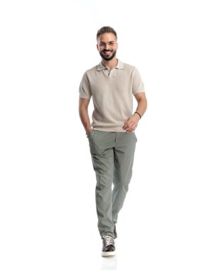 Gözlüklü, polo tişörtlü, gülümseyen ve elini cebine atarak beyaz arka plan önünde yürüyen mutlu bir adam.