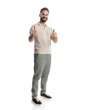 Gözlüklü çekici genç adam gülümsüyor ve beyaz arka planda dikilirken başparmak hareketi yapıyor.