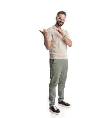 Polo tişörtlü, mutlu, sıradan bir adamın beyaz arka planda dikilirken parmaklarını arkasını göstermesi ve gülümsemesi.