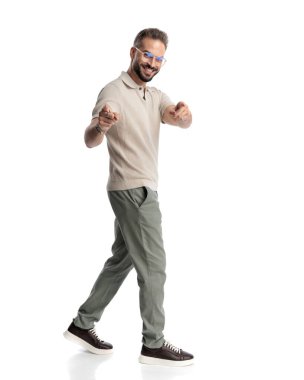 Gözlüklü, beyaz arka planda parmaklarını öne doğru gösteren ve gülümseyen sıradan genç adamın yan görüntüsü.