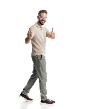 Gözlüklü mutlu genç adamın yan görünüşü. Baş parmak hareketi yapıyor ve beyaz arka planda yürüyor.