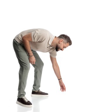 Genç, sıradan bir adam, gözlükleriyle eğiliyor, yerden bir şey alıyor ve beyaz arka planda gülümsüyor.