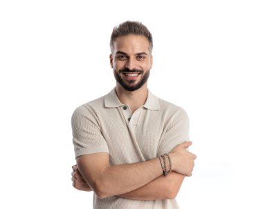 Polo tişörtlü seksi genç adam mutlu, gülümsüyor ve beyaz arka planda kollarını kavuşturuyor.