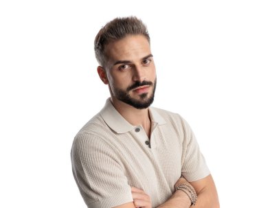 Polo tişörtlü genç bir adam beyaz arkaplanın önünde dikilirken kollarını kavuşturuyor.