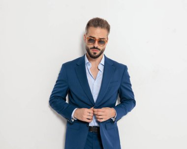 Güneş gözlüklü, lacivert takım elbiseli ve gri arka planda ileriye bakan seksi, zarif bir adam.