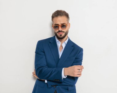 Güneş gözlüklü havalı, olgun iş adamı kollarını kavuşturup gri arka planda ileriye bakıyor.