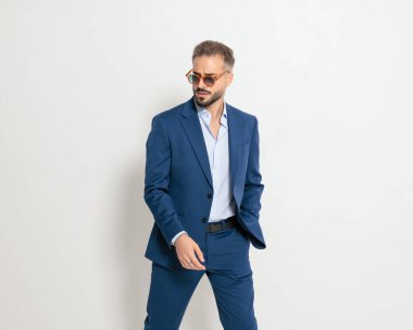 Güneş gözlüklü, zarif, olgun bir iş adamı. Gri arka planda elleri ceplerinde poz veriyor.