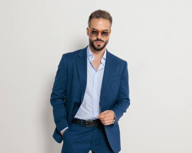 Güneş gözlüklü, lacivert takım elbiseli, cebinde el ele tutuşan ve gri arka planda duran zarif, olgun bir iş adamı.