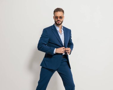 Güneş gözlüklü kibirli bir adamın portresi lacivert takım elbise ayarlıyor ve gri arka planda havalı bir poz veriyor.