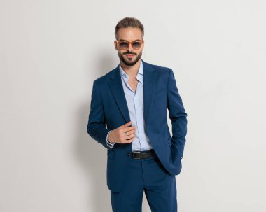 Mutlu, güneş gözlüklü, lacivert takım elbiseli ve gri arka planda gülümseyen zarif bir adamın portresi.