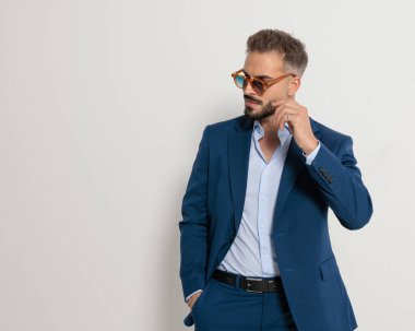 Lacivert takımlı, zarif, olgun bir iş adamı. Güneş gözlüğü takıyor ve gri arka planda el ele tutuşurken yanına bakıyor.