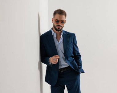 Güneş gözlüklü, havalı, olgun bir adam. Gri arka plandaki duvara yaslanıyor.