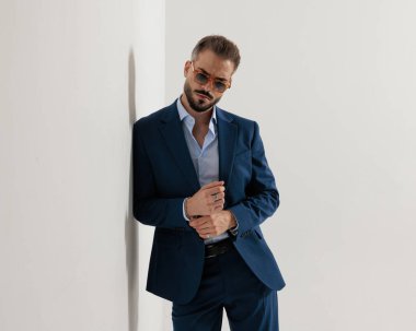 Donanma mavisi takım elbiseli havalı sakallı adam duvara yaslanıyor ve gri arka planda kollarını düzeltiyor.