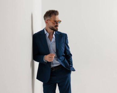 Lacivert takım elbiseli zarif genç bir işadamı, güneş gözlüğü takıyor, duvara yaslanıyor ve gri arka planda el ele tutuşuyor.