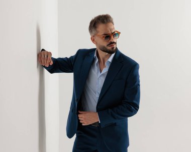 Güneş gözlüklü zarif bir iş adamı elini duvara yaslıyor ve gri arkaplanın önüne bakıyor.