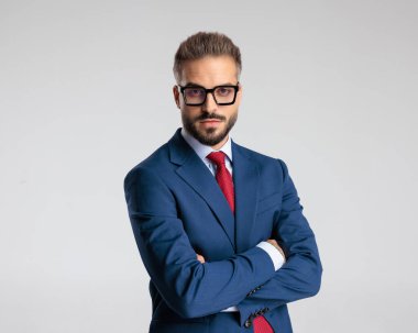 Gözlüklü, kollarını kavuşturup gri arkaplanın önünde duran ciddi olgun bir adamın portresi.