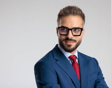 Siyah gözlüklü, lacivert takım elbiseli çekici ve zarif bir adam gri arka planda gülümsüyor.