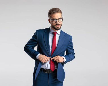 Gri arka planda dikilirken takım elbisesinin düğmelerini açmaya çalışan gözlüklü seksi genç iş adamı.