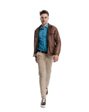 Kahverengi deri ceketli, beyaz arka planda yürüyen ve gülümseyen yakışıklı genç adamın tüm vücudu.