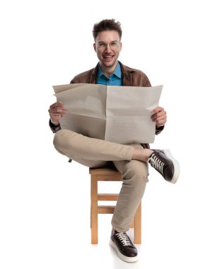 Gözlüklü heyecanlı genç adam gazete okurken gülüyor ve beyaz arka planda oturuyor.