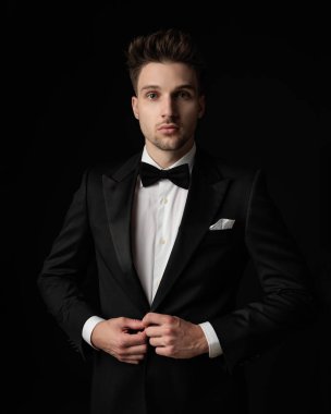 Siyah arka planda dikilirken mavi gözlü çekici adam siyah smokin giyip kapatıyor.