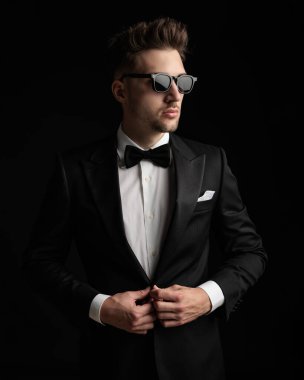 Güneş gözlüklü yakışıklı damat gözlerini kaçırıyor ve siyah arka planda siyah smokin ayarlıyor.