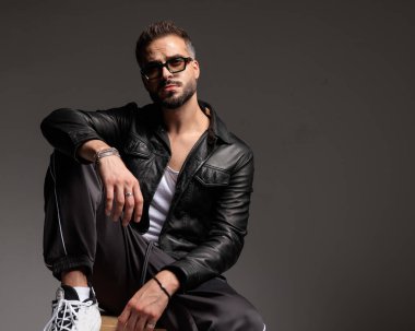 Siyah deri ceketli, dizinde dirseği olan ve kahverengi arka planda oturan çekici seksi bir adam.