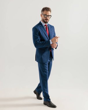 Lacivert takım elbiseli seksi sakallı adam gri arka planda kollarını düzeltiyor.