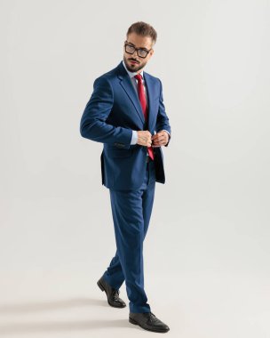 Gözlüklü, lacivert takım elbiseli, gri arkaplanın önünde yürüyen ve yana bakan zarif bir adamın yan görüntüsü.