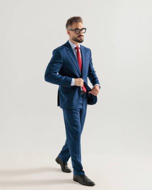 Lacivert takımlı yakışıklı, zarif iş adamı gri arka planda yürürken başka tarafa bakıyor ve takım elbise tamir ediyor.