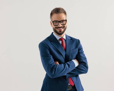 Lacivert gözlüklü genç işadamı kollarını kavuşturup gri arka planda gülümsüyor.