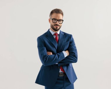 Gözlüklü, kollarını kavuşturup gri arka planda ileriye bakan zarif seksi bir adamın portresi.