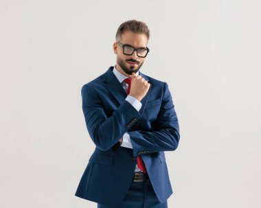 Lacivert takımlı düşünceli genç işadamı kollarını gri arkaplan önünde kavuştururken çenesine dokunup düşünüyor.
