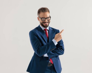 Lacivert takım elbiseli mutlu genç işadamı gülümsüyor, kollarını kavuşturuyor ve gri arka planda parmaklarını sağa sola gösteriyor.