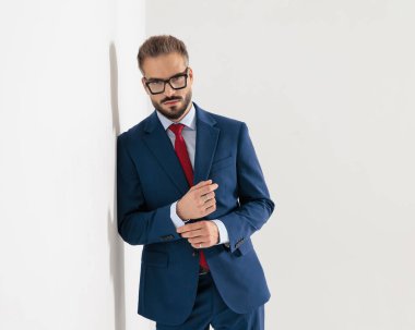 Lacivert takımlı gözlüklü seksi genç işadamı gri arka planda bir duvara yaslanırken kollarını düzeltiyor.