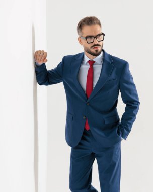 Lacivert takım elbiseli, kendine güvenen zarif bir adam, yan tarafa bakıyor, el ele tutuşuyor ve bir kolunu gri arka planda duvara yaslıyor.