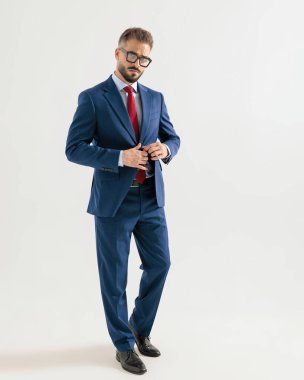 Gözlüklü seksi sakallı adam lacivert takımı kapatıyor ve gri arka planda yürüyor.