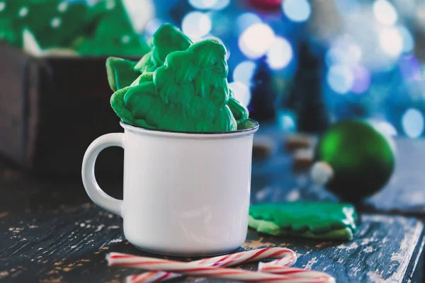 霜降りのクリスマスツリーの形のクッキーやビスケットは 近くにキャンディー缶がある白いコーヒーカップの中に緑色のアイシングがあります 前景と背景がぼやけた選択的焦点 — ストック写真