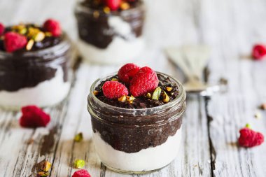 Beyaz ahşap arka planda cam bir kavanozda ahududu ve şam fıstıklı siyah çikolatalı puding. Hindistan cevizi kremalı olsun. Sağlıklı yiyecekler. Boşluğu kopyala.