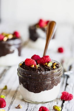 Beyaz ahşap arka planda cam bir kavanozda ahududu ve şam fıstıklı siyah çikolatalı puding. Hindistan cevizi kremalı olsun. Sağlıklı yiyecekler. Boşluğu kopyala.