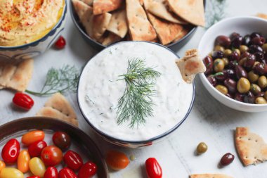 Taze domatesler, zeytinler ve humus ile çevrili bir tabak tzatziki sosu ve pide ekmeğinin beyaz kırsal bir masa üzerindeki manzarası..