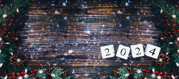 Stock image New Year's 2024 wood calendar blocks banner. Christmas tree lights, pine branches, red winter berries and snow over wooden table background. Top view, flat lay with copy space available.