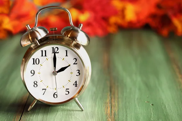 Set your clocks back with this alarm clock with colorful fall leaves over a green rustic wooden table. Daylight saving time concept. Selective focus with blurred background.