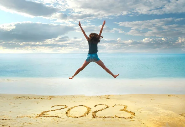 Yeni yıl 2023 kumsalda yazılmış, mutlu kız elleri havada plajda zıplıyor..