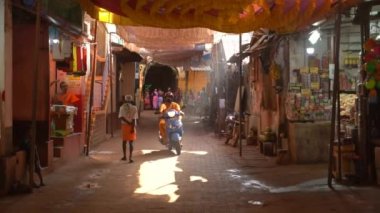 Gokarna, Hindistan - 21 Şubat 2023 - Gokarna 'da sabah, insanlar gün ışığıyla aydınlanmış bir cadde boyunca yürüyorlar. Geleneksel Shaivite turuncu elbiseli adam scooter kullanıyor..
